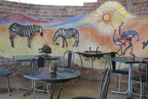 “2 of Harare’s well-known artist, Helen Lieros and Arthur Azevedo combined to provide this beautiful mural. Helen provided the background to Arthur’s amazing metal animals in relief.
He also created the crocodile, and the python on the coffee shop floor.
Lyn Barrie donated a striking painting of Wild Dogs which adorns one of the walls in the main section of the coffee shop”
